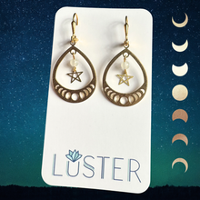 Load image into Gallery viewer, Gold teardrop earrings with star and moon phases on a Luster branded card against a starry night sky background.