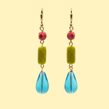 Load image into Gallery viewer, A pair of handmade drop earrings featuring gold-tone hardware, a faceted pink bead, a textured olive green cylindrical bead, and a translucent blue teardrop-shaped bead, set against a solid pale yellow background.