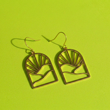 Load image into Gallery viewer, A pair of gold-tone dangle earrings with a minimalist arch design featuring a sunburst and mountain motif, displayed on a bright lime green background.