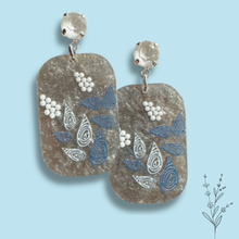Load image into Gallery viewer, A pair of handmade dangle earrings featuring textured, rounded rectangular acrylic pendants in a silvery gray hue, decorated with blue and white leaf and berry patterns, suspended from white faceted crystal studs on a light blue background.