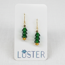 Load image into Gallery viewer, A pair of gold-tone and green drop earrings shaped like Christmas trees, displayed on a white brand card labeled 'LUSTER' against a light textured fabric background.
