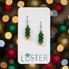 Load image into Gallery viewer, A pair of dangling earrings shaped like small green Christmas trees with gold tree toppers, displayed on a white Luster brand card against a white fabric background.

