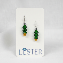 Load image into Gallery viewer, A pair of dangling earrings shaped like small green Christmas trees with silver star tops and gold tree trunks, displayed on a white Luster brand card against a light gray background.
