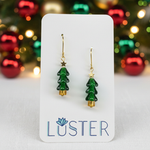 Load image into Gallery viewer, A pair of gold-tone and green enamel Christmas tree drop earrings displayed on a white Luster brand card, with a blurred festive holiday background of red and green ornaments.
