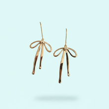 Load image into Gallery viewer, A pair of gold dangling earrings shaped like stylized bows, shown against a solid pale blue background.
