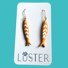 Load image into Gallery viewer, Orange fish-shaped earrings on a Luster branded card against a teal background
