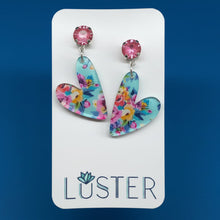 Load image into Gallery viewer, A pair of floral whimsical heart pendants on pink crystal studs, set on royal blue background.
