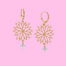 Load image into Gallery viewer, A pair of gold leverback earrings with a cutout sunburst flower design and a small ice blue faceted stone dangle, shown against a solid pink background.
