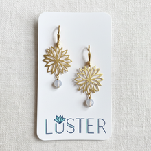 Load image into Gallery viewer, A pair of gold dangle earrings with cutout floral starburst designs and small icy blue stone drops, displayed on a white Luster brand card against a textured off-white linen background.
