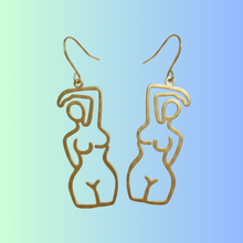 Load image into Gallery viewer, A pair of gold-tone wire cutout dangle earrings shaped like abstract minimalist female figures, shown against a soft blue-to-green gradient background.
