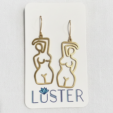Load image into Gallery viewer, A pair of gold-tone wire cutout dangle earrings shaped like abstract female figures, displayed on a white branded card that reads LUSTER.
