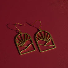 Load image into Gallery viewer, A pair of gold drop earrings with a sunburst and landscape silhouette design against a dark red background.
