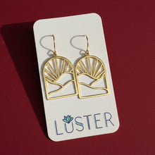 Load image into Gallery viewer, A pair of gold geometric dangle earrings displayed on a white Luster brand card against a dark red background.
