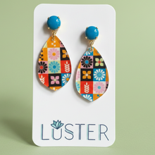 Load image into Gallery viewer, A pair of colorful patchwork teardrop dangle earrings with bright blue top beads, displayed on a white Luster brand card against a soft sage green background.

