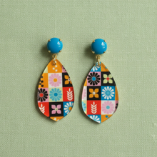 Load image into Gallery viewer, A pair of teardrop-shaped dangle earrings with a colorful retro floral patchwork pattern, suspended from bright blue round studs, displayed on a textured sage green background.
