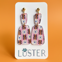 Load image into Gallery viewer, A pair of statement drop earrings with a pink background and a mid-century modern floral pattern in orange, black, white, and blue, displayed on a white Luster brand card against a solid orange background.
