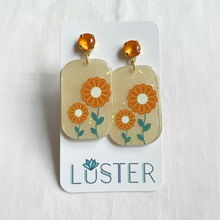 Load image into Gallery viewer, A pair of dangle earrings displayed on a white Luster brand card. The earrings feature round amber-orange faceted studs and rectangular gold-toned dangles decorated with orange flowers, white centers, and green stems on a glittery gold background.
