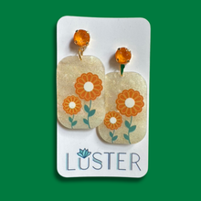 Load image into Gallery viewer, A pair of rectangular drop earrings with orange flower designs and green stems on a glittery beige background, displayed on a white card labeled 'LUSTER' against a solid green background.
