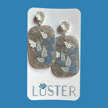 Load image into Gallery viewer, A pair of handmade dangle earrings featuring textured, rounded rectangular acrylic pendants in a silvery gray hue, decorated with blue and white leaf and berry patterns, suspended from white faceted crystal studs on a blue background.
