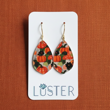 Load image into Gallery viewer, A pair of teardrop-shaped dangle earrings with a colorful pumpkin pattern, displayed on a white Luster brand card against a textured rust-orange background.
