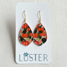 Load image into Gallery viewer, Teardrop-shaped dangle earrings with an autumn pumpkin pattern, displayed on a white Luster brand card against a textured beige fabric background.
