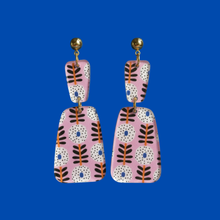 Load image into Gallery viewer, A pair of bohemian-style drop earrings with pink, white, black, and orange patterns, featuring gold-tone posts, set against a solid blue background.
