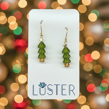 Load image into Gallery viewer, A pair of gold-tone dangle earrings with green tree-shaped charms, displayed on a white Luster brand card against a light gray textured background.

