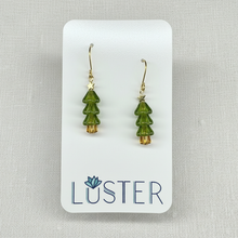 Load image into Gallery viewer, A pair of gold-tone dangle earrings with green tree-shaped charms, displayed on a white Luster brand card against a light gray textured background.
