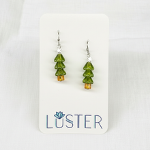 Load image into Gallery viewer, A pair of dangling earrings shaped like small green Christmas trees with amber-orange bases and silver hooks, displayed on a white Luster brand card against a light fabric background.
